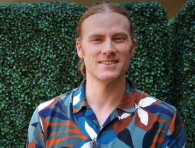 Jesse Gleaton smiling in a colorful shirt in front of a green hedge.