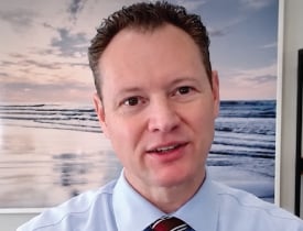 Merrill Taylor wearing a blue shirt and striped tie in front of an ocean picture.