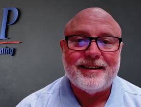 William Spar smiling in glasses and a light blue shirt against a dark wall.